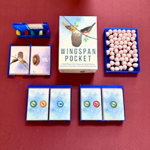 May include: A Wingspan Pocket board game box featuring an illustration of birds in a nest, accompanied by blue plastic trays containing decks of cards, small colorful wooden tokens, and a collection of light pink speckled egg-shaped game pieces.