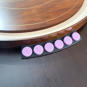 May include: A close-up of a wooden lazy susan with a black holder containing six lavender-colored circular pieces. The lazy susan has a dark brown finish with lighter wood accents. The holder is curved to fit the edge of the lazy susan.
