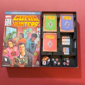 May include: A board game called "Comic Hunters" with a colorful box featuring comic book characters. The game includes a variety of components, including cards, tokens, and a game board. The box is open to reveal the game's contents.