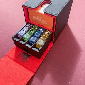 May include: A black dice box with a red interior, holding several multi-colored dice. The box is open, revealing a compartment with a gold-colored card that reads "Tanglewoods Gold". The box is on a red surface.