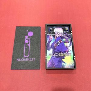 May include: A black card game box and card deck with the word "ALCHEMIST" on the box and cards. The box has a purple graphic of a test tube. The card deck features a colorful illustration of a character in purple clothing.
