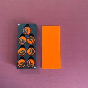May include: A black box with eight orange circular game pieces featuring a wolf silhouette. The pieces are arranged in a grid pattern. A separate orange rectangular piece is also included.