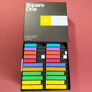 May include: A board game titled "Square One" with a black box and colorful game pieces. The box lid features the game's name, a yellow and white design, and the text "Another chapter in the PROJECT L Universe."
