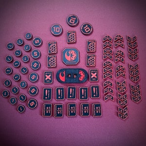 May include: A collection of red and black game pieces with various numbers and symbols. The pieces are arranged in a grid pattern on a pink surface. The pieces include numbers, letters, and symbols such as "X", "+", and "!".
