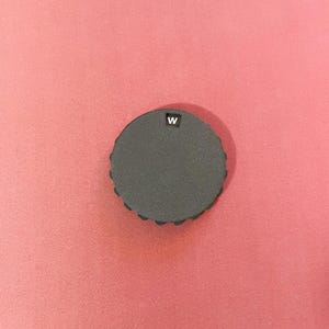 May include: A dark gray, circular object with a textured edge and the letter "W" in white. The object is set against a pink background.