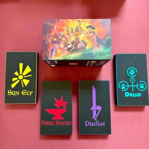 May include: A rectangular game box featuring a fantasy illustration of four characters, surrounded by four black rectangular foam inserts. Each insert displays a colored icon and text: yellow Sun Elf, red Forge Master, purple Duelist, and teal Druid.