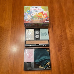 May include: An open board game box set on a wooden surface. The box lid features a colorful illustration with the text "Flamecraft Duels Deluxe Edition." Inside are various game components, including cards, dice, and a green velvet drawstring bag.