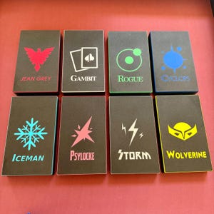 May include: Eight black card boxes with character names and symbols. The boxes feature the names Jean Grey, Gambit, Rogue, Cyclops, Iceman, Psylocke, Storm, and Wolverine. Each box has a unique color scheme and graphic design.