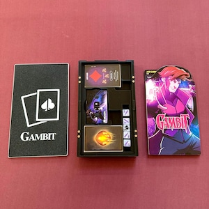 May include: A Gambit board game set with a black box containing cards and game pieces. The set includes a black card with the word "Gambit" and a spade symbol, and a card featuring a character illustration.