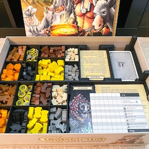 May include: A wooden game organizer with compartments for storing game pieces. The organizer has a white background with black compartments. The compartments contain various game pieces, including wooden figures, dice, and cards. The organizer is labeled "A Game of Farmers".