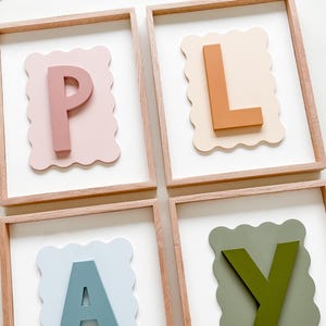 May include: Four framed wooden wall art pieces with a scalloped border. Each frame features a different color letter, spelling out the word "PLAY". The letters are pink, orange, blue, and green.
