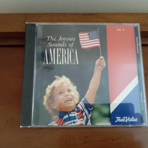 May include: A CD case titled The Joyous Sounds of America Vol. II featuring a child with curly hair holding a small American flag. The cover design includes a black background, a red and blue side panel with a white diagonal stripe, and the True Value logo.