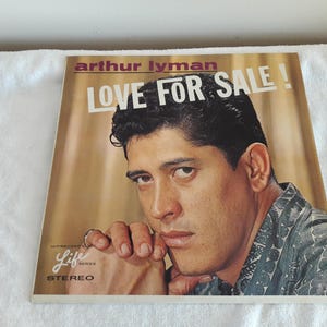 May include: Vintage vinyl record album cover for Arthur Lyman's "Love For Sale." The cover features a portrait of a man with dark hair and a patterned shirt. The album title is in large white letters.