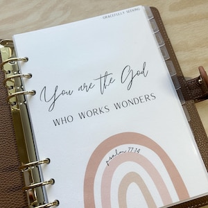 以下が含まれることがあります： 白いプランナーページに「You are the God who works wonders」というテキストと、その下に「psalm 77:14」というテキストが書かれた虹のグラフィックがあります。プランナーは茶色の革製で、金色のリングが付いています。