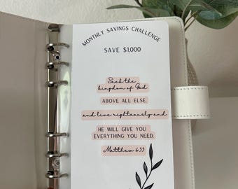 May include: A white binder with a card that reads "MONTHLY SAVINGS CHALLENGE SAVE $1,000." The card also has text that says "Seek the kingdom of God above all else, and live righteously and He will give you everything you need. Matthew 6:33."