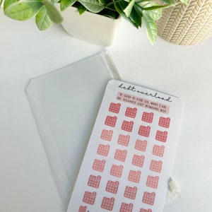 May include: A sheet of stickers with 28 calendar icons, each with a dollar sign inside. The stickers are in shades of pink and red. The text "debt overload" is at the top of the sheet. The text "IF GOD IS FOR US, WHO CAN BE AGAINST US? ROMANS 8:31" is below the icons. The text "X 28 Icons = Goals" is at the bottom of the sheet, along with the text "GRACEFULLY SEEKING".