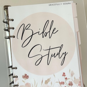Puede incluir: Un archivador de estudio bíblico con un diseño circular rosa claro y las palabras "Bible Study" en elegante escritura negra. El archivador presenta ilustraciones florales en acuarela en la parte inferior y el texto "Gracefully Seeking" en la parte superior. El archivador se mantiene abierto con anillos de metal plateado.