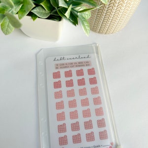 May include: A clear plastic pouch with a white card inside. The card has a pink and white design with the text "debt overload" and "If God is for us, who can be against us? Romans 8:31". There are 28 pink and white calendar icons with a dollar sign on each icon. The text "X 28 Icons = Goals =" is written on the card. The card also has the text "GRACEFULLY SEEKING".