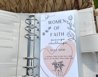 May include: A white planner with a clear plastic pocket containing a printable savings challenge card. The card features a heart design with the text "Women of Faith Savings Challenge" and "Save $400". The card also includes a quote from Proverbs 31:30, "A woman who fears the Lord shall be praised." The card is from Gracefully Seeking.
