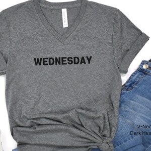 May include: Dark heather grey V-neck t-shirt with the word "WEDNESDAY" printed in black on the front.