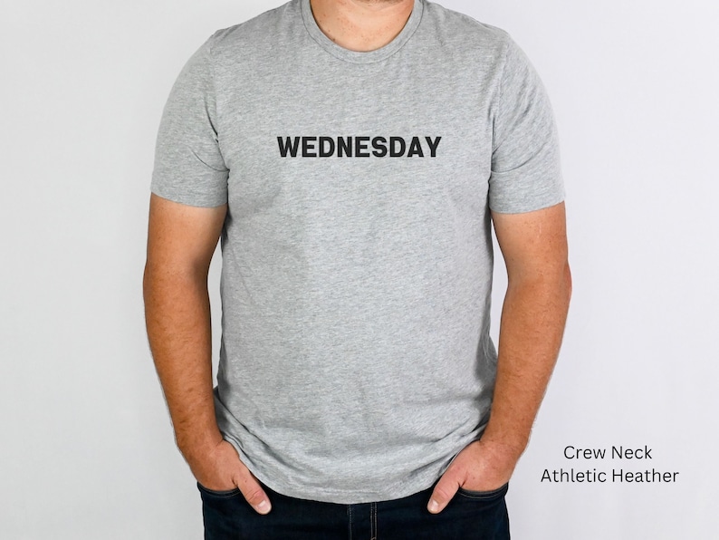 May include: A gray crew neck t-shirt with the word "WEDNESDAY" printed in black on the front. The shirt is made of athletic heather fabric.