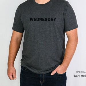 May include: A dark heather grey crew neck t-shirt with the word "WEDNESDAY" printed in black on the front.