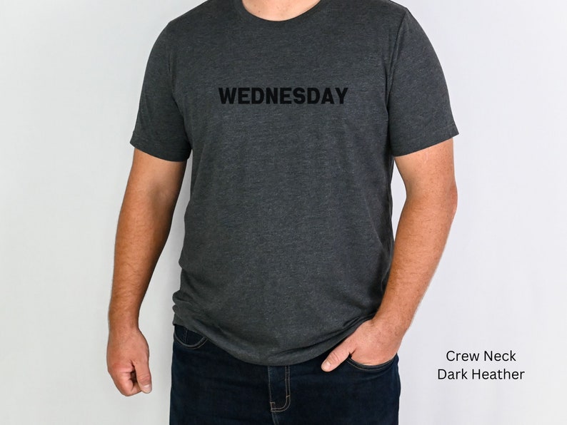 May include: A dark heather grey crew neck t-shirt with the word "WEDNESDAY" printed in black on the front.