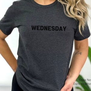May include: A dark heather gray crew neck t-shirt with the word "WEDNESDAY" printed in black on the front.