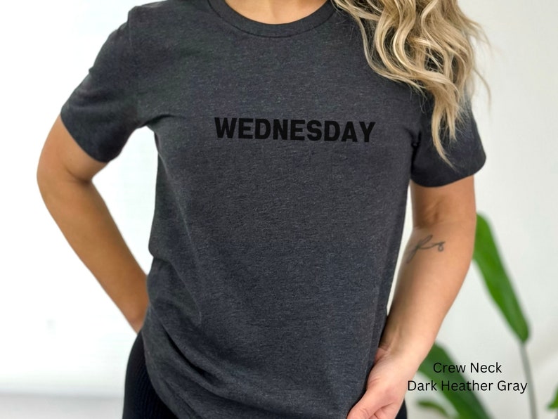 May include: A dark heather gray crew neck t-shirt with the word "WEDNESDAY" printed in black on the front.