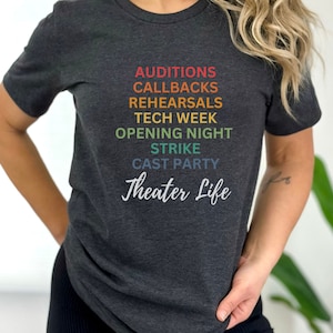 May include: A dark heather grey t-shirt with a rainbow design featuring the words "Auditions, Callbacks, Rehearsals, Tech Week, Opening Night, Strike, Cast Party, Theater Life".