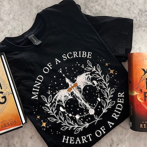 May include: A black t-shirt with a white design featuring a dragon, stars, and the text "Mind of a Scribe Heart of a Rider". Two fantasy novels are also pictured, one with a red cover and the text "Iron Flame" and the other with an orange cover and the text "Urthwing".