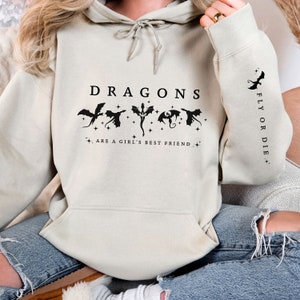 May include: A light beige hoodie with a black graphic print of dragons and the text "DRAGONS ARE A GIRL'S BEST FRIEND." The right sleeve has the text "FLY OR DIE." printed vertically.