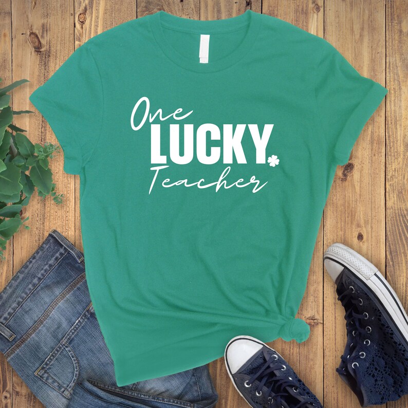 Pu&ograve; includere: Una maglietta turchese con la scritta "One LUCKY Teacher" in bianco e in stampatello. Un piccolo quadrifoglio bianco &egrave; accanto alla parola "LUCKY". La maglietta &egrave; annodata in basso. Sono visibili jeans blu e una sneaker blu navy.