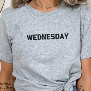 May include: A gray crew neck t-shirt with the word "WEDNESDAY" printed in black on the front.