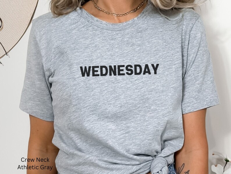 May include: A gray crew neck t-shirt with the word "WEDNESDAY" printed in black on the front.