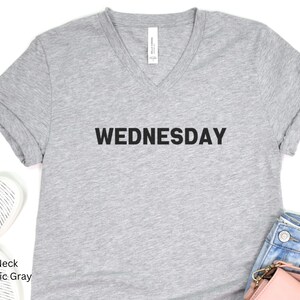 May include: A gray V-neck t-shirt with the word "WEDNESDAY" printed in black on the front.