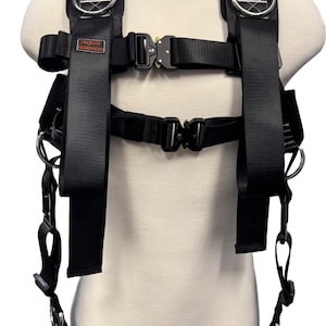 May include: Black squat harness with two adjustable straps and four D-rings. The harness has the text "SQUAT HARNESS" printed on the front strap.