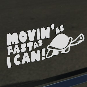 Peut inclure: Autocollant en vinyle blanc avec le texte "MOVIN' AS FAST AS I CAN!" et une illustration de tortue de dessin animé.
