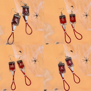 May include: Novelty earrings in the shape of blood bags, with silver hooks. Each bag is labeled with a blood type: B, O, AB, or A. The bags are filled with red liquid and have red tubing.