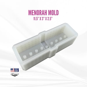 Op de afbeelding: Een witte Menorah-mal, afmetingen 29,2 cm x 7,6 cm x 6,4 cm. De mal heeft een rechthoekige vorm met verhoogde secties en kleine, cilindrische uitsteeksels. De tekst "MENORAH MOLD" wordt boven de afmetingen weergegeven. De afbeelding bevat ook een "Made in USA"-logo.