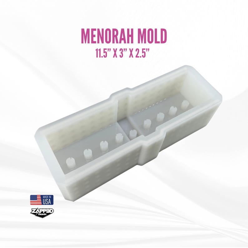 Op de afbeelding: Een witte Menorah-mal, afmetingen 29,2 cm x 7,6 cm x 6,4 cm. De mal heeft een rechthoekige vorm met verhoogde secties en kleine, cilindrische uitsteeksels. De tekst "MENORAH MOLD" wordt boven de afmetingen weergegeven. De afbeelding bevat ook een "Made in USA"-logo.
