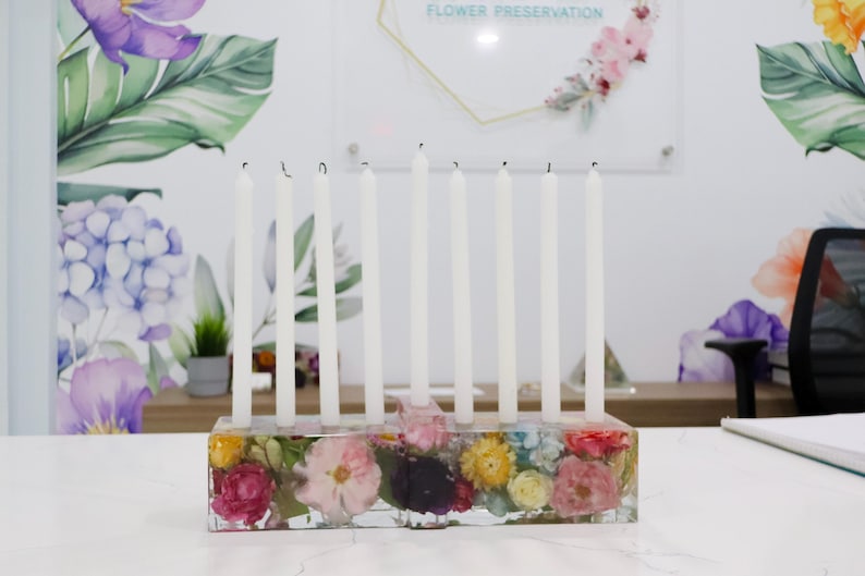 Op de afbeelding: Een heldere hars kaarsenhouder met kleurrijke gedroogde bloemen ingebed in de basis. Negen witte kaarsen zijn in de bovenkant geplaatst. De achtergrond heeft bloemenbehang en een bord met "FLOWER PRESERVATION".