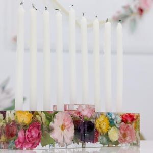 Op de afbeelding: Een decoratieve kandelaar met negen witte kaarsen. De basis is een helder harsblok gevuld met kleurrijke gedroogde bloemen. De bloemen omvatten roze, gele en rode bloemen, waardoor een levendige en artistieke weergave ontstaat.