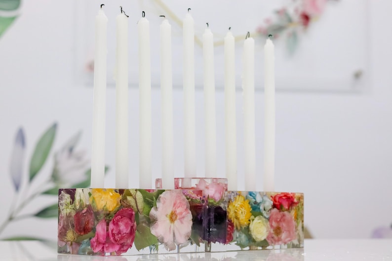 Op de afbeelding: Een decoratieve kandelaar met negen witte kaarsen. De basis is een helder harsblok gevuld met kleurrijke gedroogde bloemen. De bloemen omvatten roze, gele en rode bloemen, waardoor een levendige en artistieke weergave ontstaat.