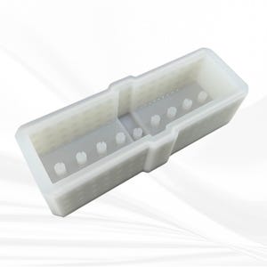 Op de afbeelding: Een witte rechthoekige plastic mal met een verdeeld interieur. De mal heeft een reeks kleine, verhoogde pinnen en een gestructureerd oppervlak. Het ontwerp suggereert dat het bedoeld is voor het maken van meerdere kleine objecten.