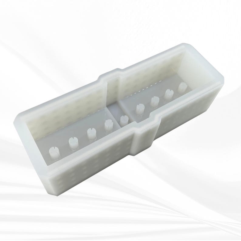 Op de afbeelding: Een witte rechthoekige plastic mal met een verdeeld interieur. De mal heeft een reeks kleine, verhoogde pinnen en een gestructureerd oppervlak. Het ontwerp suggereert dat het bedoeld is voor het maken van meerdere kleine objecten.