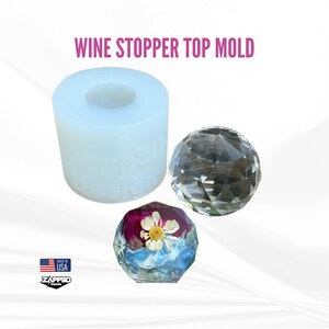 May include: A clear silicone mold for making wine bottle stoppers. The mold is shaped like a round, faceted gem. A finished wine stopper made with the mold is shown next to the mold. The stopper is filled with resin and dried flowers.