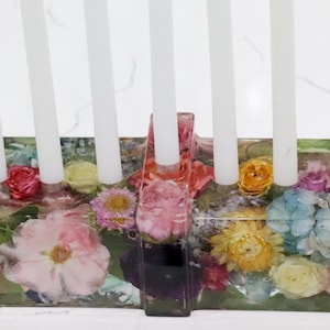 Op de afbeelding: Een bloemen menorah met witte kaarsen. De basis heeft een rechthoekige vorm en is gevuld met kleurrijke gedroogde bloemen, waaronder rozen en madeliefjes. De bloemen zijn ingekapseld in een heldere hars.