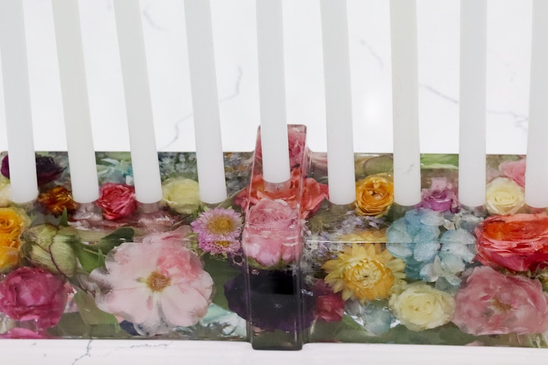 Op de afbeelding: Een bloemen menorah met witte kaarsen. De basis heeft een rechthoekige vorm en is gevuld met kleurrijke gedroogde bloemen, waaronder rozen en madeliefjes. De bloemen zijn ingekapseld in een heldere hars.