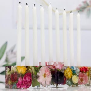 Op de afbeelding: Een bloemenkaarsenhouder van hars met negen witte kaarsen. De rechthoekige basis is gevuld met kleurrijke bloemen. De kaarsen zijn wit en zijn opgebrand. Een decoratief stuk voor de woondecoratie.